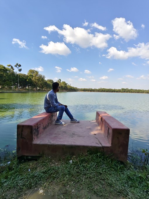 The image shows a man sitting alone on a bench near a body of water, possibly a lake or pond. The scene appears to be set during the day with a clear blue sky above him. There are no other people visible in the picture, which adds to the peaceful atmosphere. The photo is related to the post because it features an image of the man sitting on the bench and the surrounding environment, including trees and water.