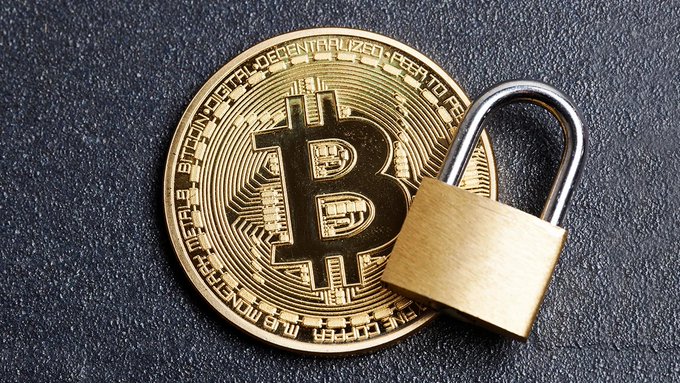A gold physical Bitcoin coin with 'VIRES IN NUMERIS' and 'DIGITAL DECENTRALIZED PEER TO PEER' engraved around the edge rests on a dark surface, secured with a brass padlock through its center.