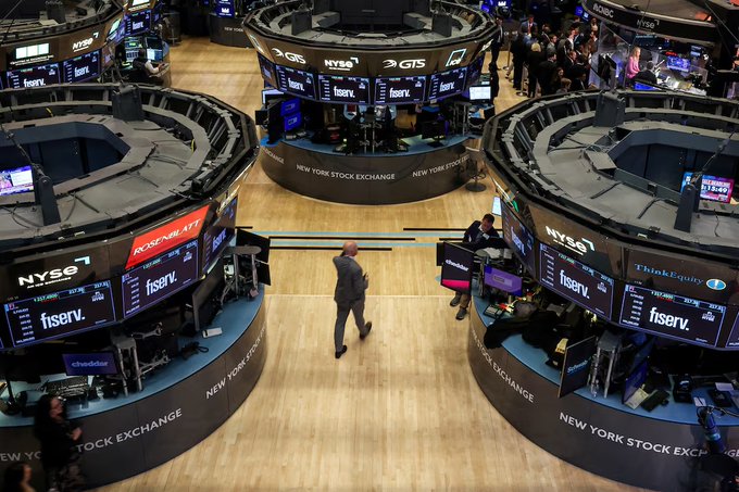 An overhead view shows the trading floor of the New York Stock Exchange, where traders and monitors display "NYSE," "GTS," "ROSENBLATT," "ThinkEquity," and "fiserv" branding on circular trading posts. One trader in a dark suit walks across the polished wood floor between the desks. The image relates to the post by illustrating a traditional stock exchange venue like the NYSE, where Nasdaq's $100 million investment in Payward, the parent of crypto exchange Kraken, aims to bring tokenized stock trading into such conventional financial market infrastructure.