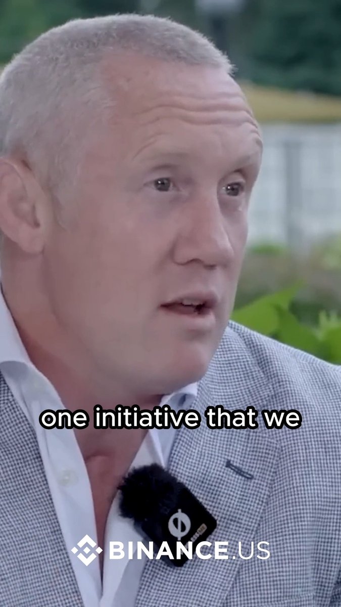 A man with gray hair, wearing a checkered blazer over a white shirt with a microphone clipped to his lapel, appears mid-speech in what looks like an outdoor interview setting. The "BINANCE.US" logo is displayed at the bottom along with the subtitle "one initiative that we," tying into the post about Binance.US's 0% maker fees spot trade promotion.