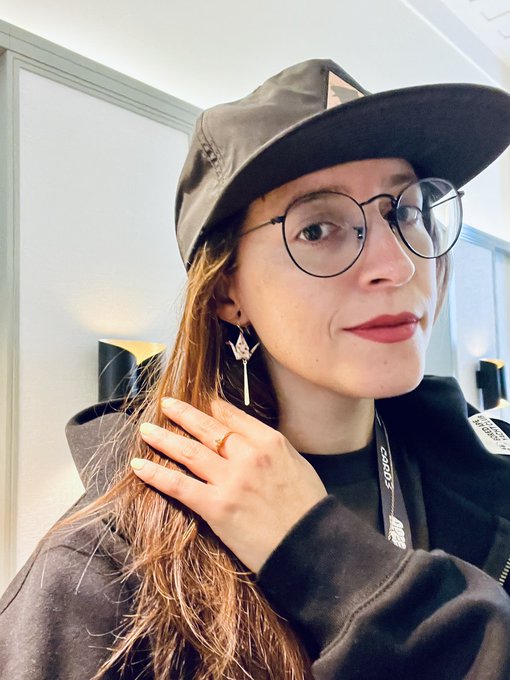 A woman wearing a cap, round glasses, and distinctive earrings poses for a selfie while touching her hair.