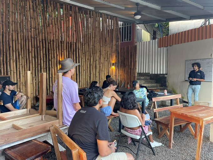A group of people attending what appears to be a presentation or class in an open-air venue with bamboo walls, where one person stands by a whiteboard.