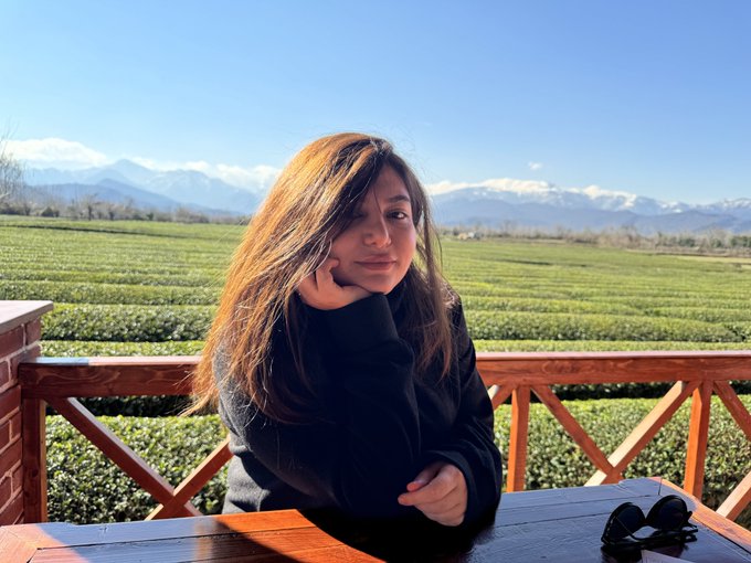 A woman sits at a wooden table on a balcony overlooking lush green tea fields and snow-capped mountains under a bright blue sky.