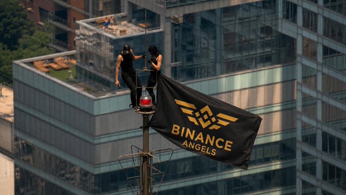 Two performers perched atop a ship mast unfurl a black banner emblazoned with the "BINANCE ANGELS" logo, bringing dramatic flair to the urban skyline. The bold, high-altitude display visually echoes the post's message that "Angels' love always wins."