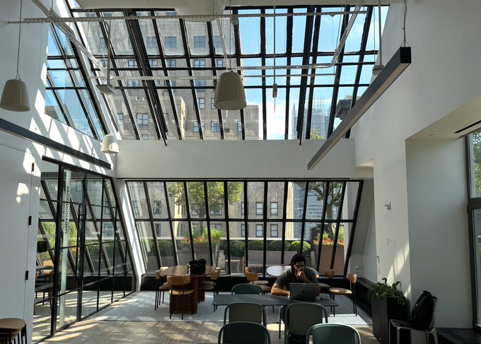 A man works on a laptop in a sunlit, modern lounge-style coworking space with floor-to-ceiling windows and glass roofing, where empty chairs and tables await additional visitors. The bright, open environment reflects the post's invitation to join the remaining open spots at the Fireblocks NYC office co-working session.