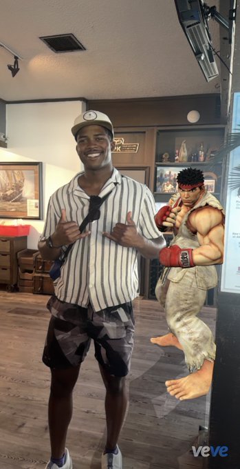 A smiling man in a striped shirt and cap poses with a "Ryu" cardboard cutout (Street Fighter) in what appears to be a themed bar or restaurant, with the "veve" logo visible in the corner, marking his welcome to the VeVe digital collectibles family.