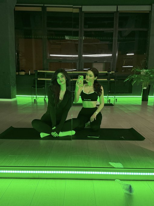 Two women in black workout attire take a mirror selfie in a dance or fitness studio with green LED lighting along the floor.