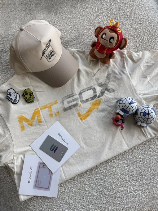 A flat lay photograph displays merchandise including a beige 'MEOW' cap, matching t-shirt, small plush toys, soccer ball figures, and white packaging on a carpet.