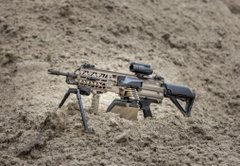 A tan and black tactical rifle with a scope, foregrip, and bipod is displayed on a dirt surface. The image visually demonstrates the post's point about normalizing the casual sharing of firearms online, highlighting the potential security risks of openly showcasing weapon details.