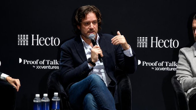 The image shows a man sitting in a chair at a conference with two other people behind him. The man has his hand raised, possibly speaking or gesturing during a presentation. There are several water bottles placed around them, indicating that they might be attending an event where refreshments are provided. The background features the logos of "HUGE" and "Profit of Talk", suggesting that this is likely a conference related to blockchain technology or cryptocurrency.