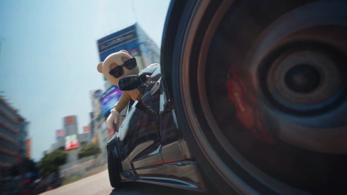 A dramatic low-angle shot captures a black sports car with a teddy bear wearing sunglasses hanging out the passenger window, speeding through a city with motion blur.