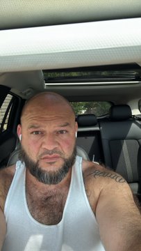 A bearded, bald man wearing a white tank top and earbuds sits inside a car with a sunroof open.