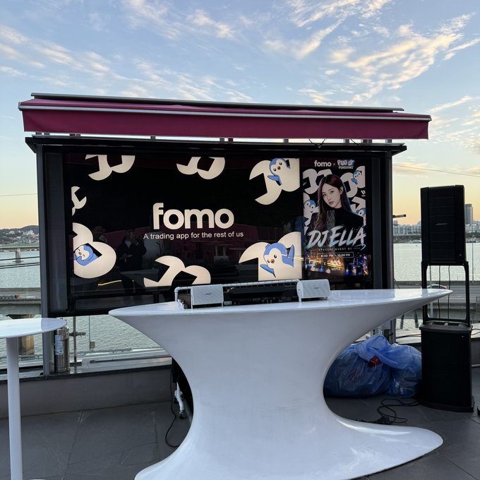 An outdoor event setup with a white cocktail table positioned in front of a digital display advertising the 'fomo' trading app with a Dizella partnership, set against a waterfront view at dusk.