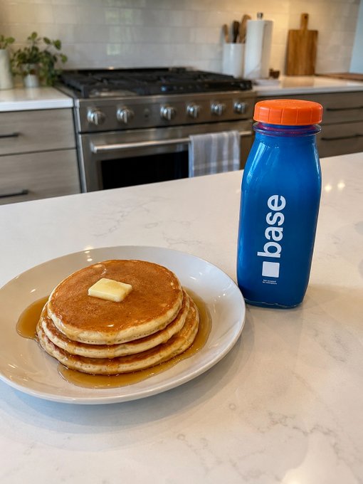 A stack of pancakes topped with a pat of butter and syrup on a white plate, sitting next to a blue 'base' branded water bottle on a marble kitchen countertop.