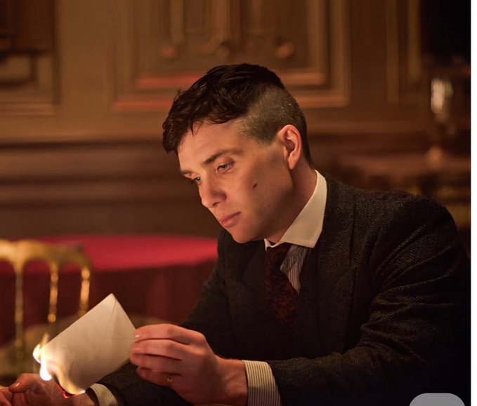 A man with dark undercut hair in a tweed jacket holds a burning letter, his face illuminated by the flame in a dimly lit room.