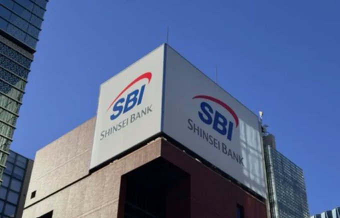 A building rooftop displays the blue and red 'SBI Shinsei Bank' logo prominently against a clear blue sky.