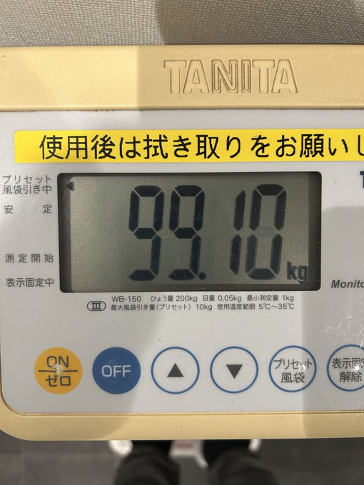 A TANITA WB-150 digital scale displays a weight reading of 99.10 kg, with a yellow sticker showing Japanese text "使用後は拭き取りをお願い" (please wipe after use).