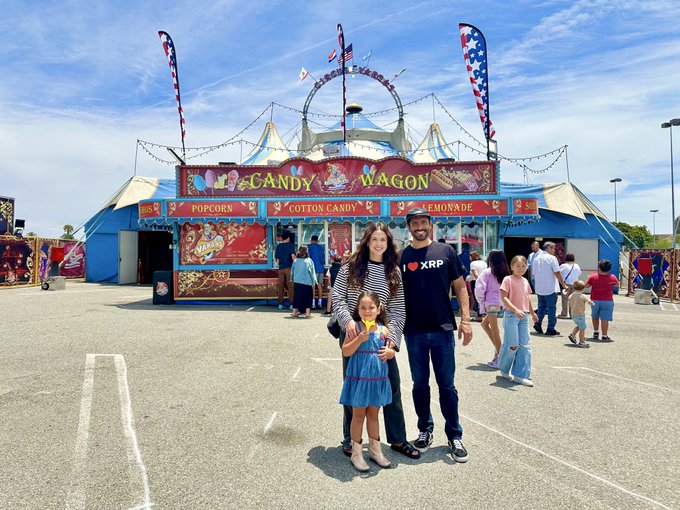 The image shows a family of three, consisting of two adults and a child, standing outside a circus tent. The man is wearing a black shirt, while the woman has her hair styled in pigtails. They are posing for a photo with smiles on their faces. In front of them, there's a food stand that sells cotton candy, adding to the festive atmosphere of the event.
