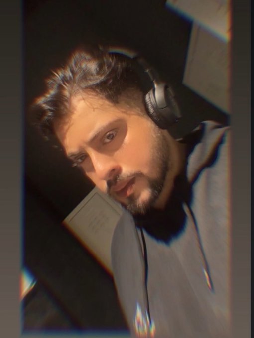 A young man with dark hair and a beard wears large black headphones, captured in a dimly lit, moody photo with a vintage filter effect.