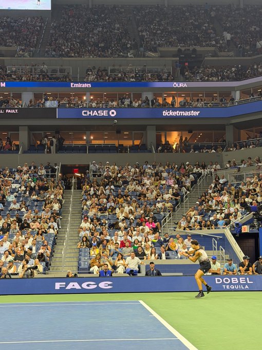 A view from the stands of a tennis player in white shorts and dark shirt hitting a shot during a match at a large stadium, likely the US Open given the Chase, Emirates, and Ticketmaster signage visible in the arena.