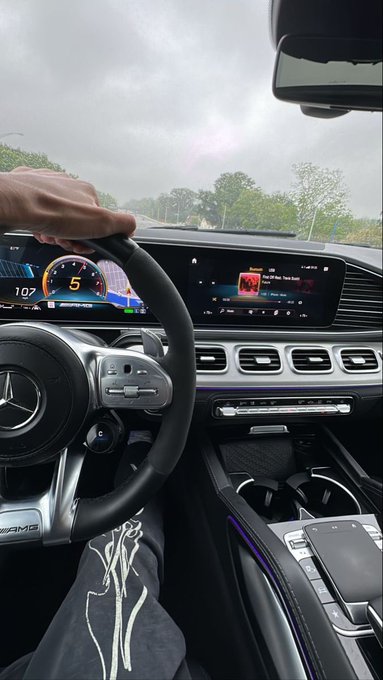 A hand grips the steering wheel of a Mercedes-Benz as the driver cruises down a tree-lined highway, with the digital dashboard displaying "107" and the infotainment screen tuned to "Pop 101 Teen Twitch." The AMG logo on the steering wheel and the sunlit road ahead embody the weekend's call to "touch grass," paired with a morning crypto shoutout to @River4fun and $River.