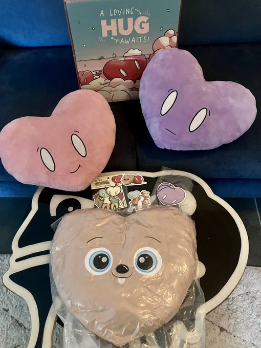 An assortment of plush toys and merchandise on a blue couch, including a 'A LOVING HUG <AWAITS>' card, a pink heart plush and purple heart plush with faces, character stickers, and a large brown heart-shaped plush with a cute face in plastic packaging.