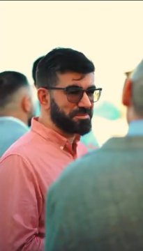A vertical portrait of a bearded man wearing glasses and a pink shirt, looking to the side in a social setting.