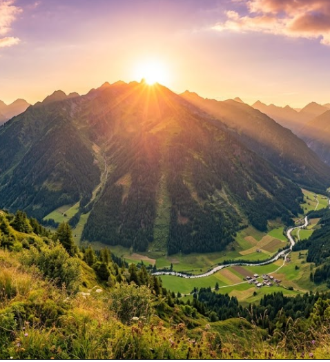 A breathtaking mountain landscape at sunset with golden sunlight beaming over the peaks and a green valley below.