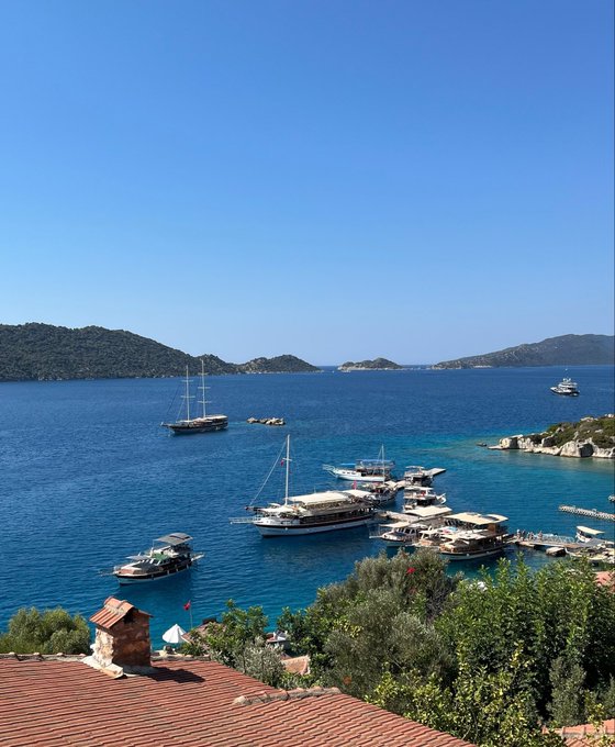 An elevated view captures a stunning Mediterranean cove with crystal-clear blue waters dotted with sailboats and yachts, surrounded by forested hills under a clear sky.