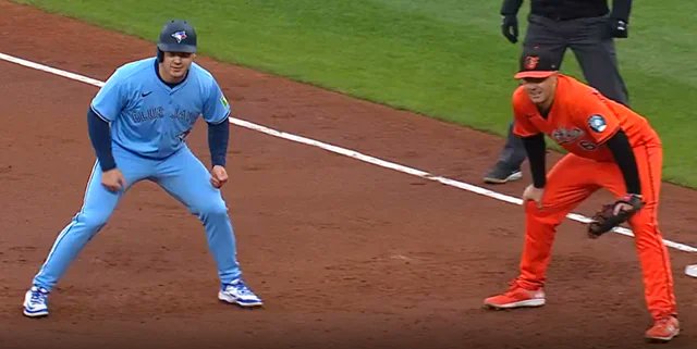 Two baseball players, one in a light blue Toronto Blue Jays uniform and the other in an orange Baltimore Orioles uniform, take their leads off a base during a game, perfectly capturing the "Orioles vs Blue Jays" matchup.