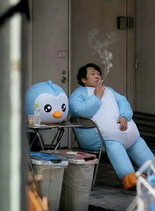 A man in an inflatable blue penguin costume smokes a cigarette while slumped against a doorway next to another inflatable penguin head and some plastic bins.
