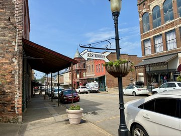 A street scene outside an antique mall with a hanging 'ANTIQUES RESTROOMS' sign, brick buildings, hanging flower baskets, and parked cars along the road.