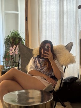 A casual mirror selfie of a young woman in a white t-shirt sitting in a butterfly chair, taking the photo with an iPhone in a bright room with plants and sheer curtains.