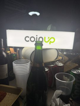 A dimly lit cluttered table holds cups, bottles, and a drum, with an illuminated "coinup" sign glowing in the background.