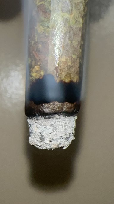The image shows a smoldering glass pipe with burnt residue and a chunk of ash at the bowl, referenced in the post as a "Pi zen" (a slang term for a piece of psilocybin). The post captions it as being "strong as shit," highlighting the perceived potency of the substance being smoked.