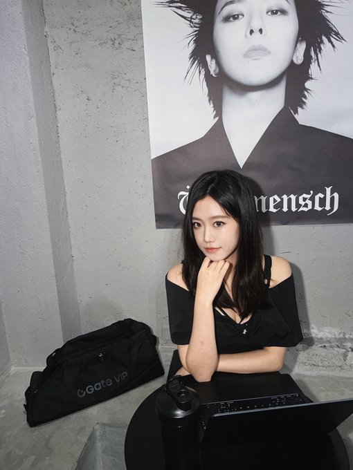 A photo of a young woman with long dark hair wearing a black off-shoulder top, seated at a table with a laptop, next to a G·Gate VIP bag, in front of a black and white poster featuring a woman's face with partial text '...mensch'.