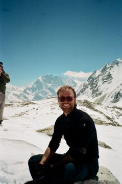 A smiling man wearing sunglasses and a dark jacket poses in the snow with majestic snow-capped Himalayan mountains towering behind him under a clear blue sky. The accompanying post references Nepal relief efforts and the @USDC ticker, suggesting this photo was taken during a journey through the Himalayan region, likely to raise awareness or support for Nepal relief initiatives via the nepalrelief.org donation platform.