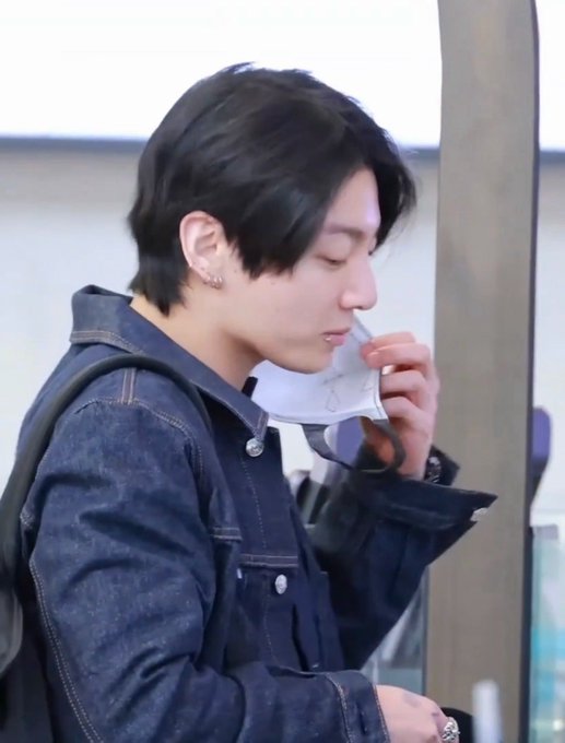 A person with dark hair, wearing a dark denim jacket and a backpack, is shown in profile holding a white face mask near their chin with their right hand. They have a small earring visible in their ear, and the background appears to be a light-colored indoor setting.
