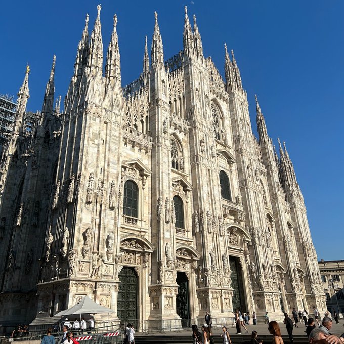 The Milan Cathedral, a Gothic masterpiece with multiple spires and statues, stands majestically against a clear blue sky, drawing the attention of tourists and locals alike.