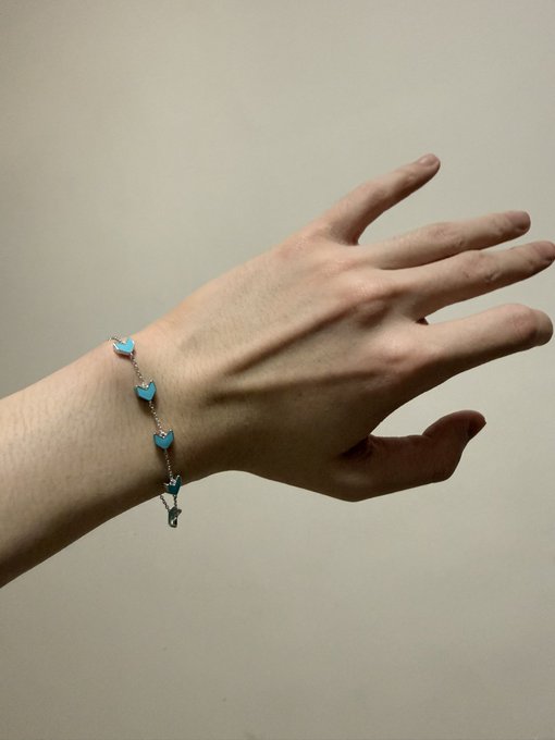 Close-up photo of a pale hand and wrist wearing a delicate bracelet with small blue heart charms and a pendant, set against a plain off-white background.