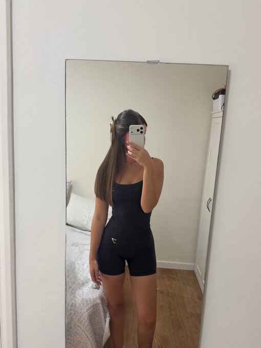 A mirror selfie of a woman in a black workout set (tank top and shorts) holding a phone that obscures her face, standing in a bedroom with a white bed and wardrobe.