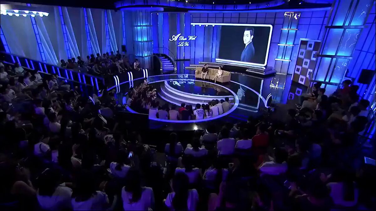 The image shows a large audience gathered around an event stage, with people sitting in rows of chairs facing a speaker who appears to be giving a speech or presentation. The stage has blue lighting that illuminates the scene, creating a captivating atmosphere for the attendees. The crowd is engaged and attentive, indicating that they are interested in what the speaker has to say.