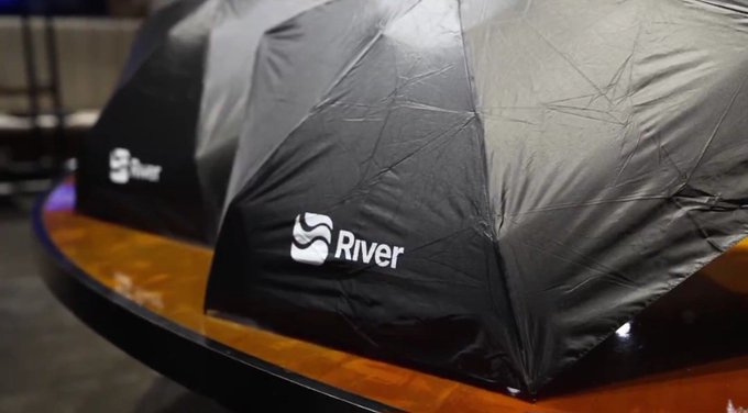The image features a black umbrella with "River" written on it, placed on top of a wooden table. There are two umbrellas in total - one larger umbrella positioned towards the left side of the frame and another smaller umbrella located more towards the center. Both umbrellas have the word "River" printed on them, indicating that they might be related to an event or gathering involving the river theme. The presence of these umbrellas suggests a possible outdoor activity or gathering taking place in a park or similar setting.