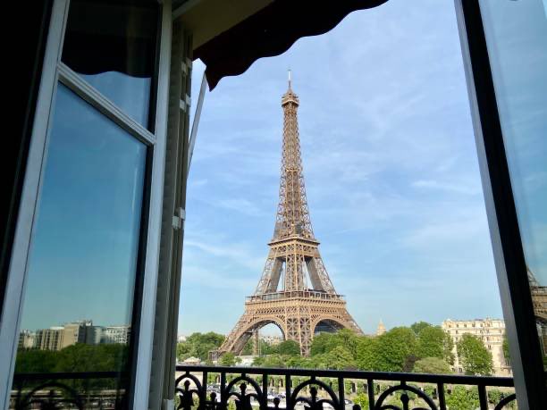 A view of the Eiffel Tower in Paris, framed by an open window and wrought-iron balcony railing with awnings above.
