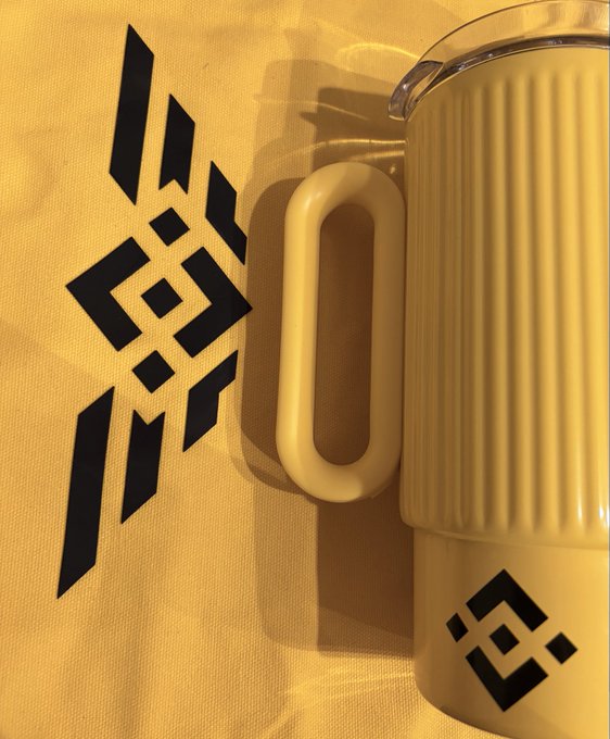 Close-up of a cream-colored ribbed mug with a black geometric logo on it, placed next to a matching yellow cloth or bag displaying the same logo.