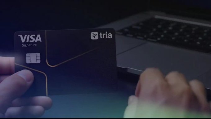 A person holds a black Visa Signature credit card branded 'tria' in front of a laptop keyboard in a dimly lit setting.