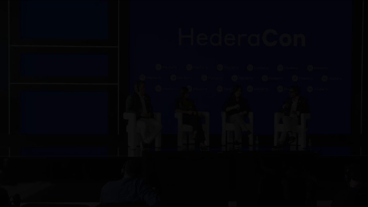 The image shows a group of people sitting around a table at a conference or event. The attendees are engaged in conversation, possibly discussing topics related to open-source software and innovation. The setting appears to be indoors with a stage visible behind them, suggesting that this gathering might be taking place at a conference or similar event.