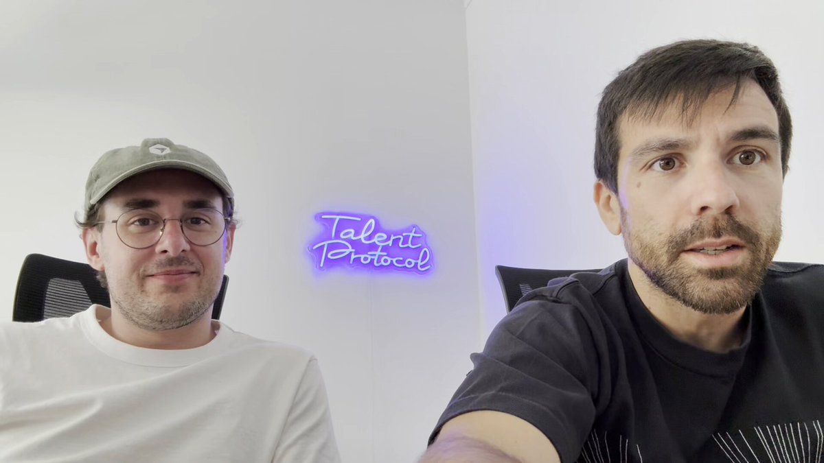 The image shows two men sitting next to each other, both wearing glasses and a cap. They are looking at the camera with smiles on their faces. The background of the image features a white wall with a neon sign that reads "TALENT PROACTIV". This suggests that they might be participating in an online event or competition related to talent development or recognition.