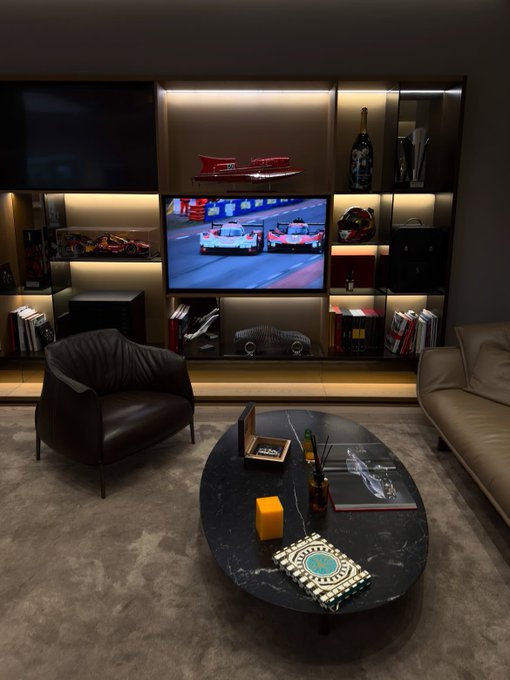 An interior view of a modern living room or den featuring a central television showing a race car video game, flanked by illuminated shelving holding car models and books, with a leather armchair and an oval coffee table in the foreground.