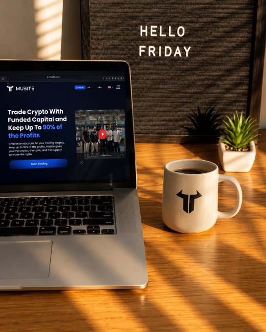 A laptop displaying the Mubite crypto trading website sits next to a branded mug on a wooden desk, with 'HELLO FRIDAY' text above.